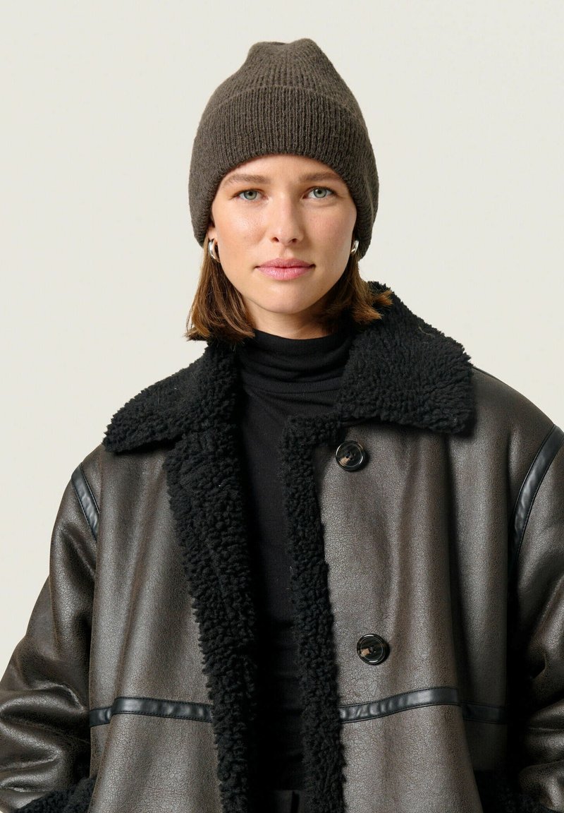 Braune Strickmütze, strukturiert, mit spitz zulaufendem Oberteil. Mit Lammfell gefütterte schwarze Lederjacke mit großem Kragen und glänzenden Knöpfen. Schwarzes Rollkragenoberteil darunter.