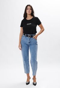 Camiseta negra con el texto "COOL", jeans de cintura alta de lavado claro y tacones negros con detalles decorativos. Diseño simple, ajuste casual.