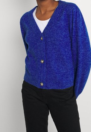 Personne portant un cardigan bleu duveteux boutonné sur un haut blanc, associé à un pantalon noir, sur un fond gris uni.