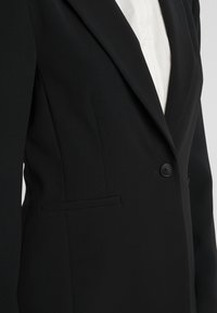 Černý blazer vyrobený z pevné tkaniny, s jednořadým zapínáním na knoflík, klopovými límci a boční kapsou. Bílá košile viditelná pod ním.