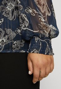 Blusa floreale navy con tessuto trasparente e motivo a strisce dorate. Polsini arricciati con dettagli elastici. Tessuto liscio e design fluido.