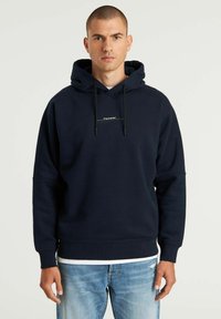 Felpa blu navy con cappuccio regolabile, caratterizzata da un logo bianco sul petto e polsini a coste. Indossata sopra jeans blu sbiaditi.