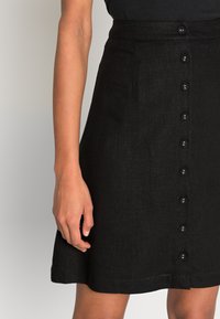 Falda negra de lino con botones, de forma acampanada, que cuenta con varios botones en la parte delantera y un dobladillo que termina por encima de la rodilla.