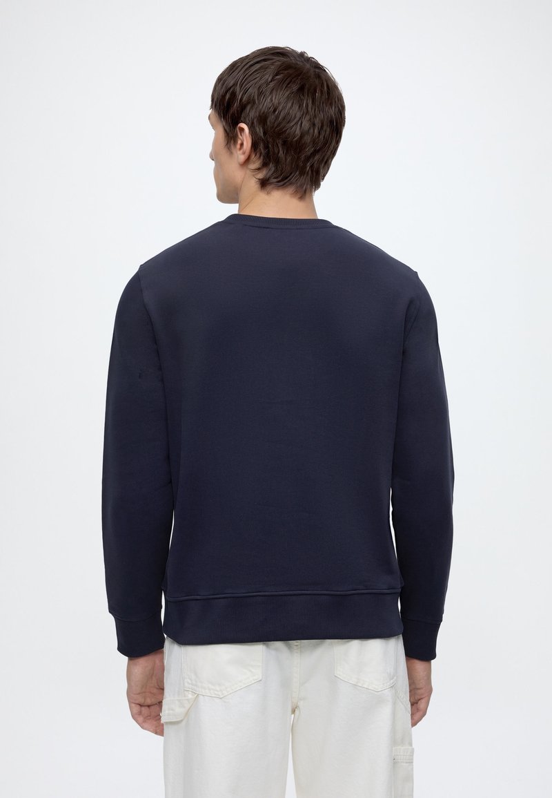 Mand iført en ensfarvet mørkeblå sweatshirt og lysebukser vist bagfra mod en ensfarvet hvid baggrund.