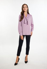 Liliowa bluzka z długimi rękawami, z detalem w postaci kokardy przy kołnierzyku oraz z zapięciem na guziki; zestawiona z czarnymi spodniami typu slim-fit i czarnymi szpilkami.