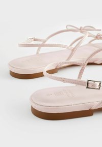 Lyserøde flade sandaler med tynde stropper og en spænde detalje, der har en glat overdel og en struktureret sål med et let brunt accent.