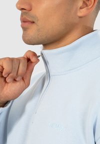 Sweat-shirt zippé bleu clair avec un col haut ; présente une fermeture éclair argentée et un logo en relief sur la poitrine en fil assorti.