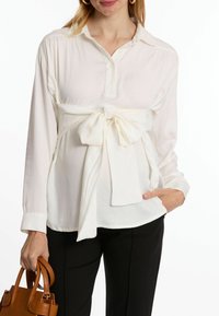 Weiße Bluse mit langen Ärmeln und Kragen, tailliert mit einem großen Schleifendetail, kombiniert mit schwarzen Hosen und einer braunen Handtasche.