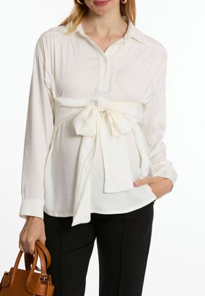 Weiße Bluse mit langen Ärmeln und Kragen, tailliert mit einem großen Schleifendetail, kombiniert mit schwarzen Hosen und einer braunen Handtasche.
