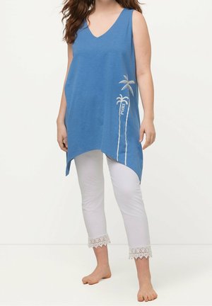 Femme portant une tunique bleue sans manches avec imprimé palmiers et des leggings blancs courts avec bordure en dentelle, debout pieds nus sur un fond uni.