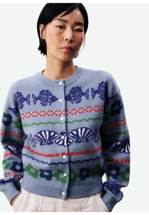 Femme portant un cardigan bleu boutonné avec des motifs de poissons, coquillages, fleurs et lignes ondulées rouges, les mains dans les poches d'un pantalon beige.