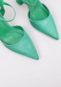 Zapatos de tacón alto de satén verde con puntera puntiaguda y correas en el tobillo, que muestran una textura suave y un tacón bloque moderado.