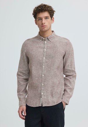 Ung mann med krøllete hår iført beige skjorte med knapper og mørke bukser, stående med en hånd i lommen mot en ensfarget bakgrunn.