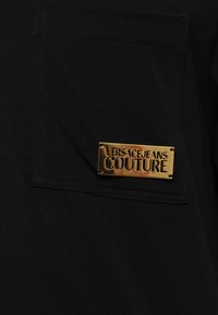 Svart bomull T-skjorte med lomme foran som har en gull rektangulær logo plate med teksten "VERSACE JEANS COUTURE." Myk tekstur, avslappet design.