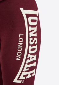 Bordeauxfarbene Leggings mit eng anliegender Passform, die ein großes weißes "Lonsdale"-Logo und den Text "London" vertikal an der Seite aufweisen. Weicher Stoff.