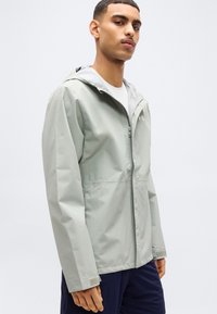 Νέος άνδρας που φοράει ανοιχτό γκρι μπουφάν με κουκούλα, φερμουάρ κλειστό πάνω από ένα λευκό πουκάμισο, σε συνδυασμό με σκουρόχρωμο παντελόνι, που στέκεται μπροστά σε απλό φόντο.