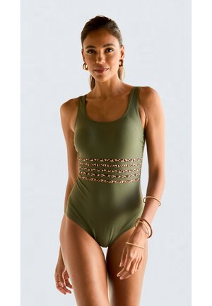 Vrouw draagt een olijfgroene badpak met luipaardprint gaasbanden in de taille, gouden creolen oorbellen en armbanden, staand tegen een witte achtergrond.
