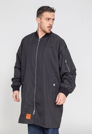 Man draagt een lange zwarte bomberjas met rits en een oranje patch, kijkt naar links tegen een effen lichte achtergrond.