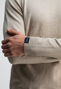 Beige Strickpullover mit gerippten Bündchen und einem Etikett am Handgelenk. Der Stoff wirkt weich und strukturiert und präsentiert ein lässiges, bequemes Design.