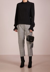 Blusa nera a maniche lunghe con polsini arricciati, abbinata a pantaloni grigi a quadri. La modella tiene una piccola borsa nera con tracolla a catena. Stivali neri con tacco.