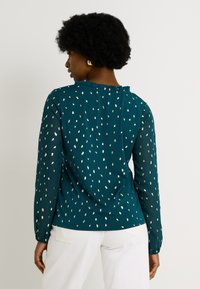 Blouse teal avec un col à volants, de longues manches en voile et des pois métalliques éparpillés, assortie à un pantalon blanc.