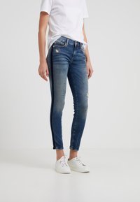 Person som bär urvuxna blå slim fit-jeans med svarta sidostråk, en vit t-shirt och vita snörskor på en enkel bakgrund.