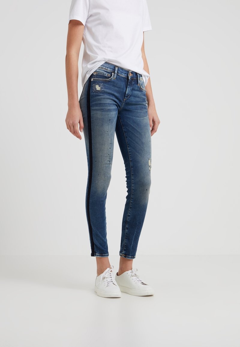 Person som bär urvuxna blå slim fit-jeans med svarta sidostråk, en vit t-shirt och vita snörskor på en enkel bakgrund.