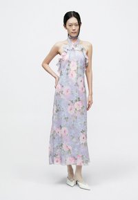 Maxi-jurk van doorschijnende lavendelkleurige stof met roze rozen en groene bladeren, voorzien van geruite bandjes en een rechte silhouet. Witte schoenen met hak.