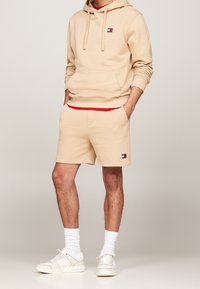 Beige katoenen hoodie met een voorzak en logo, gecombineerd met bijpassende beige shorts met een rode tailleband en logo, gedragen met witte sneakers.