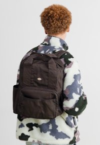 Mochila de tela marrón con un bolsillo frontal y un logo, llevada sobre una chaqueta de forro polar camuflada de color claro con un alto cuello.