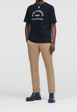 T-shirt grafica nera con testo bianco e dorato, pantaloni beige sartoriali e stivali lacciati blu navy. L'outfit presenta una vestibilità rilassata.