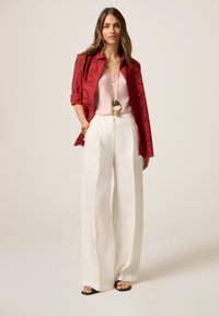 Rote bestickte Jacke über einem rosa Oberteil, kombiniert mit weißen, hoch taillierten, weiten Hosen und schwarzen Sandalen mit Akzenten. Langes, glattes Haar.