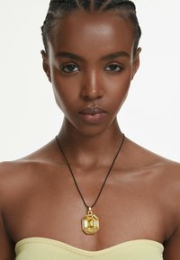 Guld octagonal vedhæng på en sort snor, båret over en gul strapless top. Vedhænget har et geometrisk design med en poleret overflade.