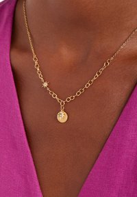 Gouden ketting met een ronde hanger met een sterontwerp en een kleine accentsteen. Draagmode tegen een dieproze stoffen achtergrond.