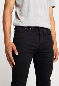 Vaqueros de denim negros con corte ajustado, que cuentan con una cremallera, cinco bolsillos y detalles de costura sutiles. Combinados con una camiseta gris de manga corta.