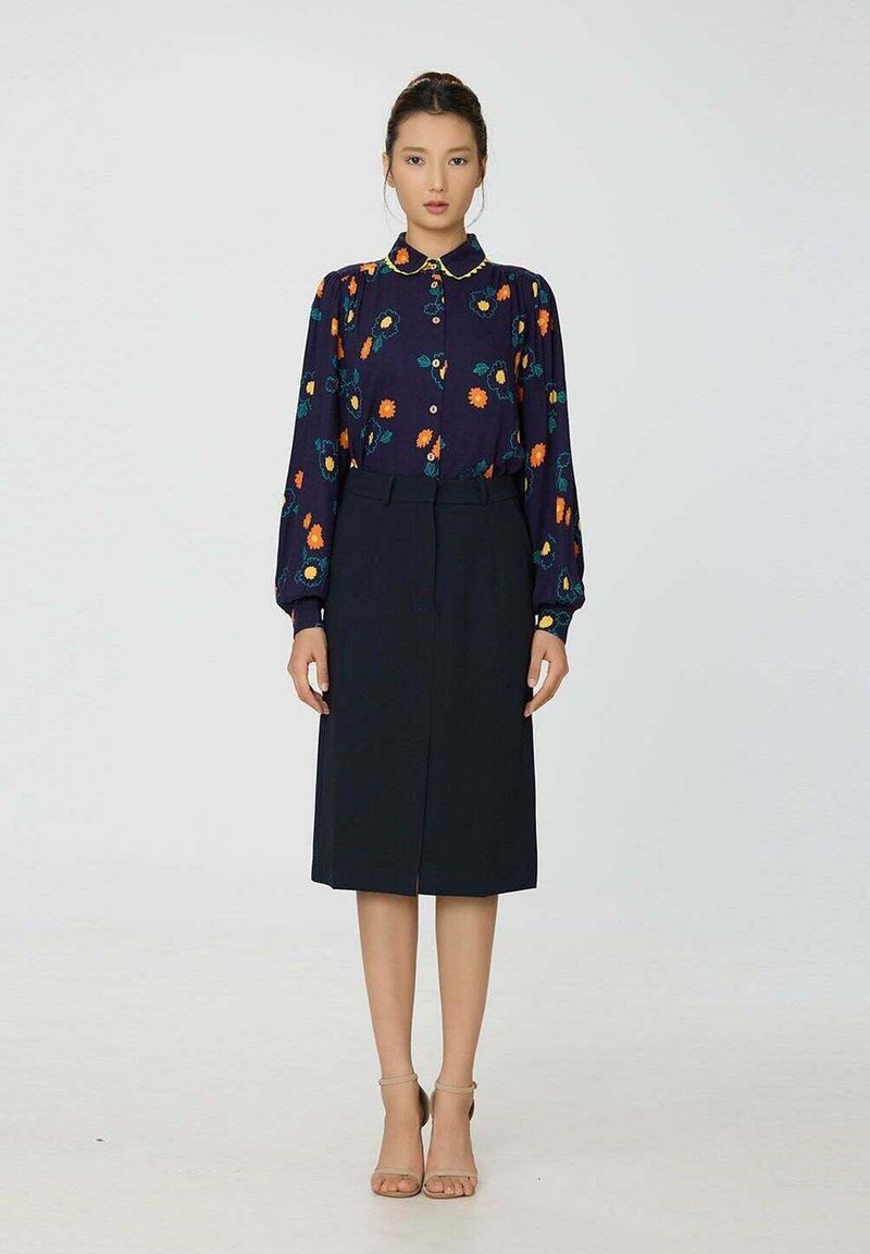 Blouse florale bleue marine avec un col jaune et des manches longues, associée à une jupe noire jusqu'aux genoux. Le mannequin se tient devant un fond uni.