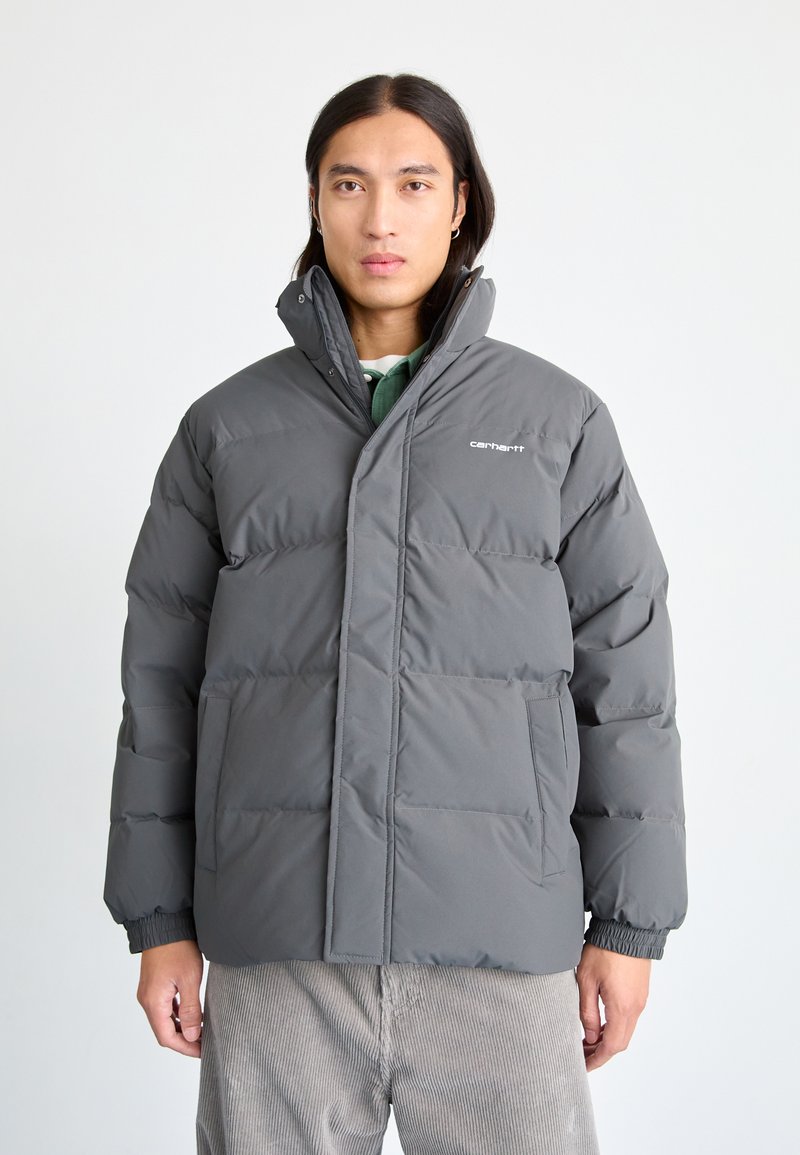 Chaqueta acolchada gris con cuello alto, cierre frontal de botones a presión, puños elásticos y dos grandes bolsillos frontales. Presenta un sutil logo de la marca.