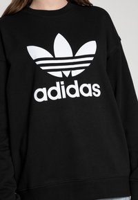 Crna dukserica od pamuka s okruglim izrezom. Sadrži veliki bijeli Adidas trefoil logotip i horizontalne pruge u dizajnu.