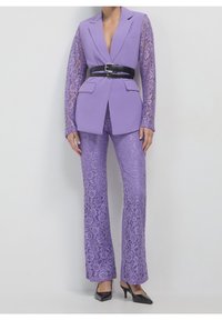 Fioletowy koronkowy garnitur z dopasowaną marynarką o fakturowanych rękawach, w zestawie z dopasowanymi koronkowymi spodniami i czarnym paskiem. Buty na wysokim obcasie.