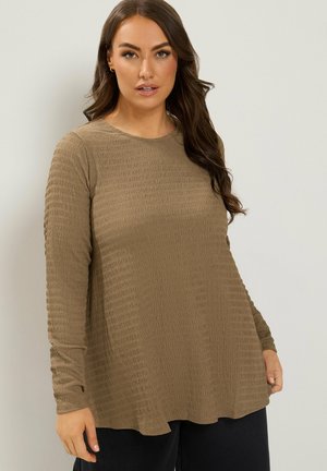 Femme aux longs cheveux bruns porte un haut marron texturé, ample à manches longues, et un pantalon noir sur un fond uni.