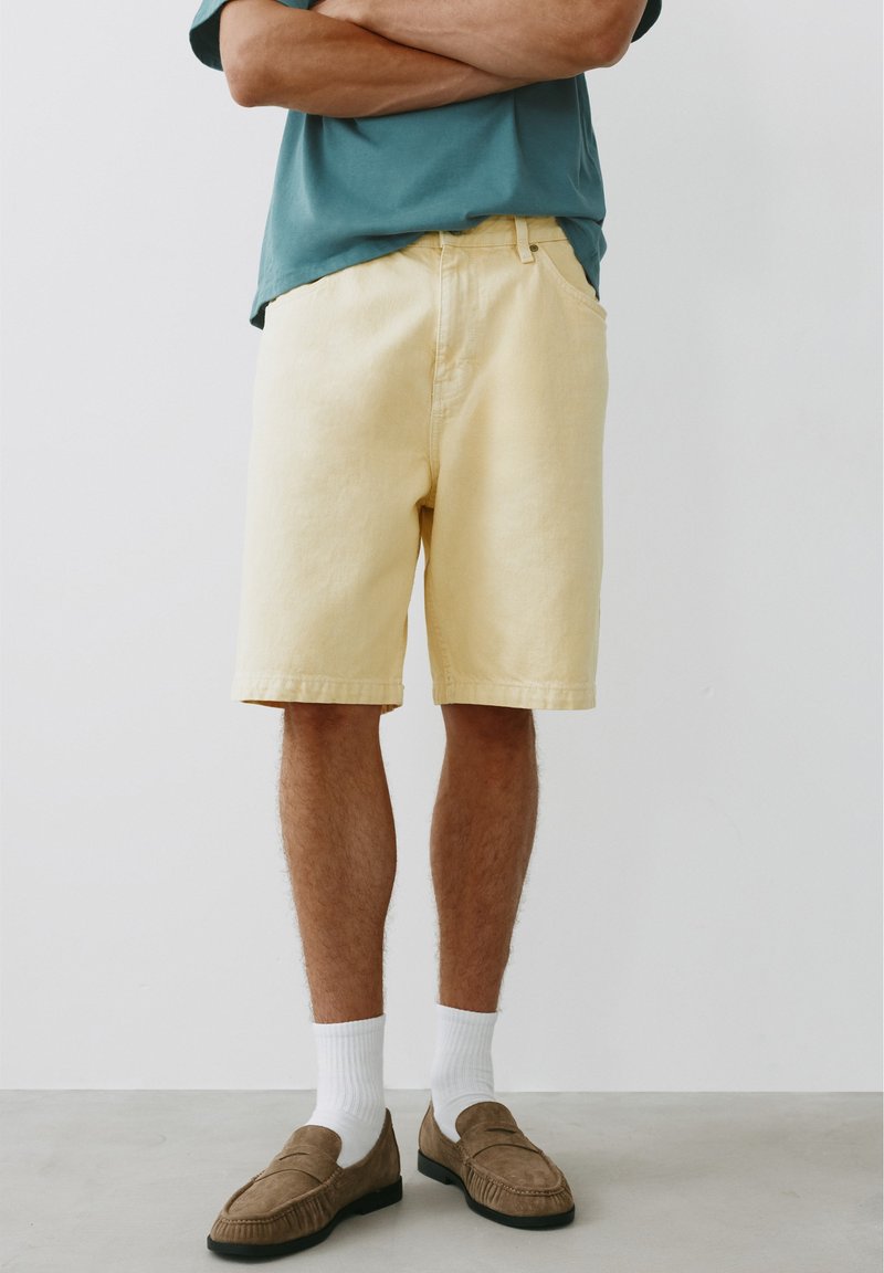 Personne portant un short jaune clair jusqu'aux genoux, des chaussettes blanches, des chaussures marron sans lacets et un t-shirt bleu sarcelle à manches courtes, bras croisés.