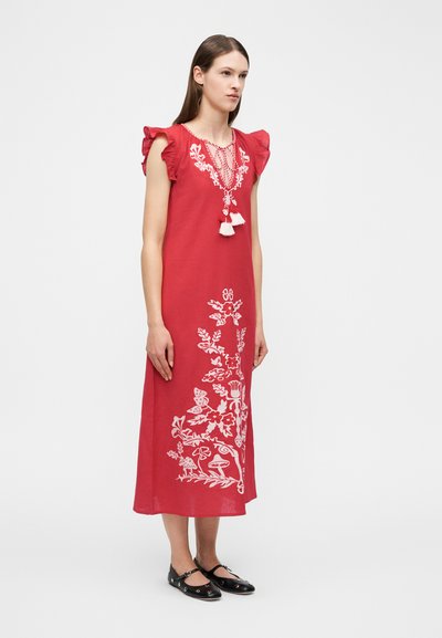 Γυναίκα με κόκκινο midi φόρεμα χωρίς μανίκια, διακοσμημένο με λευκά λουλουδένια κεντήματα, ριγωτούς ώμους, κορδόνια με φούντες και μαύρα παπούτσια Mary Jane.