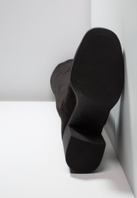 Botte de cheville en suédine noire avec une finition texturée, dotée d'un talon épais et d'une semelle en caoutchouc. Botte posée sur une surface blanche.