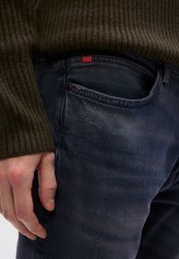 Mørkeblå denimjeans med en lett tekstur, med en rød etikett nær midjen og en frontlomme. En ribbestrikket olivengrønn genser delvis synlig.