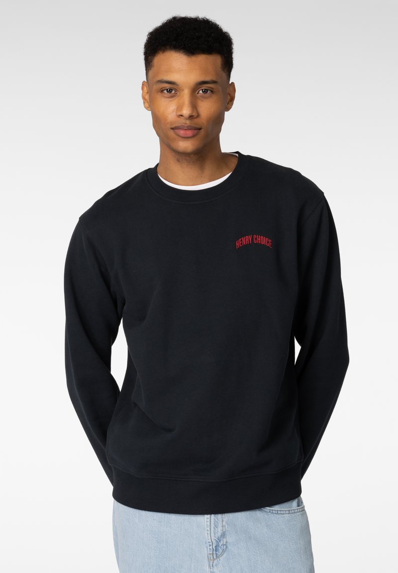 Svart hettegenser laget av bomullsblanding, med ribbestrikket rund hals, lange ermer, og rødt brodert tekst "HENRY CHOICE" på brystet.