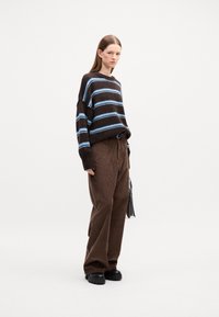 Braun gestreiften Pullover mit blauen Linien, kombiniert mit braunen Cordhosen und klobigen schwarzen Schuhen. Das Model hält eine kleine schwarze Clutch.