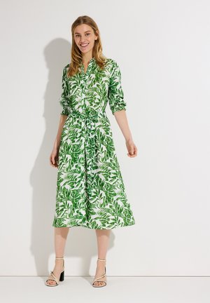 Frau lächelnd, trägt ein grün-weißes, mit Blättern gemustertes Midikleid mit Knöpfen und hochgekrempelten Ärmeln sowie beigen Sandalen mit hohen Absätzen, steht vor einer weißen Wand.