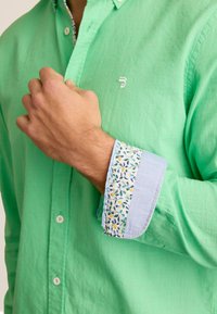 Groene linnen shirt met een button-down kraag. De manchetten hebben een bloemmotief in blauw, geel en groen op een lichtblauwe achtergrond.