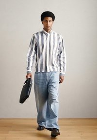 Ριγέ πουκάμισο με κουμπιά σε λευκό και γκρι, ανοιχτό μπλε τζιν και μαύρα παπούτσια. Κρατώντας μαύρη τσάντα χειρός. Φόντο ξύλινο πάτωμα.