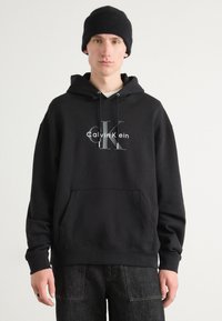 Crni pamučni dukserica s prednjim kengur džepom, s printanim Calvin Klein logotipom u sivoj boji i podesivim vezicama na kapuljači.